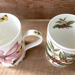 May include: Two white ceramic mugs featuring botanical illustrations. The left mug displays a large pink flower and a yellow butterfly. The right mug shows coffee beans, green leaves, and white blossoms. Both mugs have matching interior motifs.