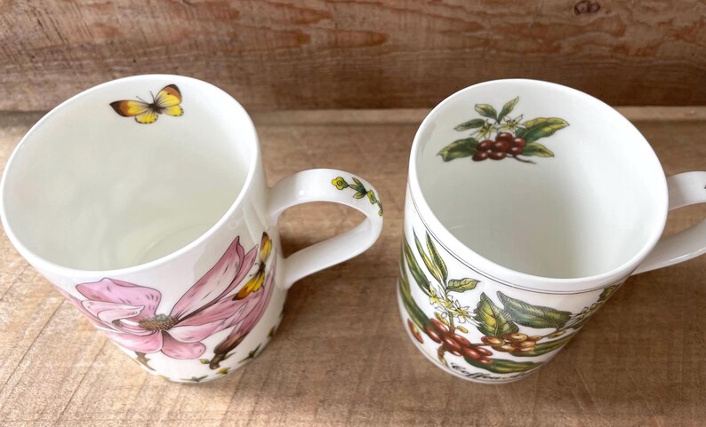 May include: Two white ceramic mugs featuring botanical illustrations. The left mug displays a large pink flower and a yellow butterfly. The right mug shows coffee beans, green leaves, and white blossoms. Both mugs have matching interior motifs.