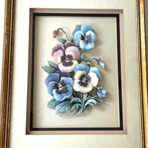 May include: A framed artwork depicting a bouquet of pansies. The flowers are in shades of blue, purple, pink, and white, with green leaves. The artwork is set against a light background, framed in gold.