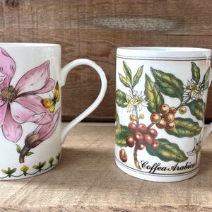 May include: Two white ceramic mugs with handles. The left mug features a large pink magnolia flower and a small yellow butterfly. The right mug displays a botanical illustration of a coffee plant with green leaves, white flowers, and red berries, labeled Coffea Arabica.