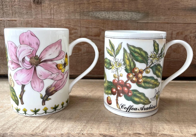 May include: Two white ceramic mugs with handles. The left mug features a large pink magnolia flower and a small yellow butterfly. The right mug displays a botanical illustration of a coffee plant with green leaves, white flowers, and red berries, labeled Coffea Arabica.