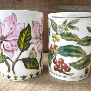 May include: Two white ceramic mugs featuring botanical illustrations. The left mug displays large pink magnolia flowers, green leaves, and a small butterfly. The right mug features coffee plant branches with green leaves, white flowers, and red coffee cherries.