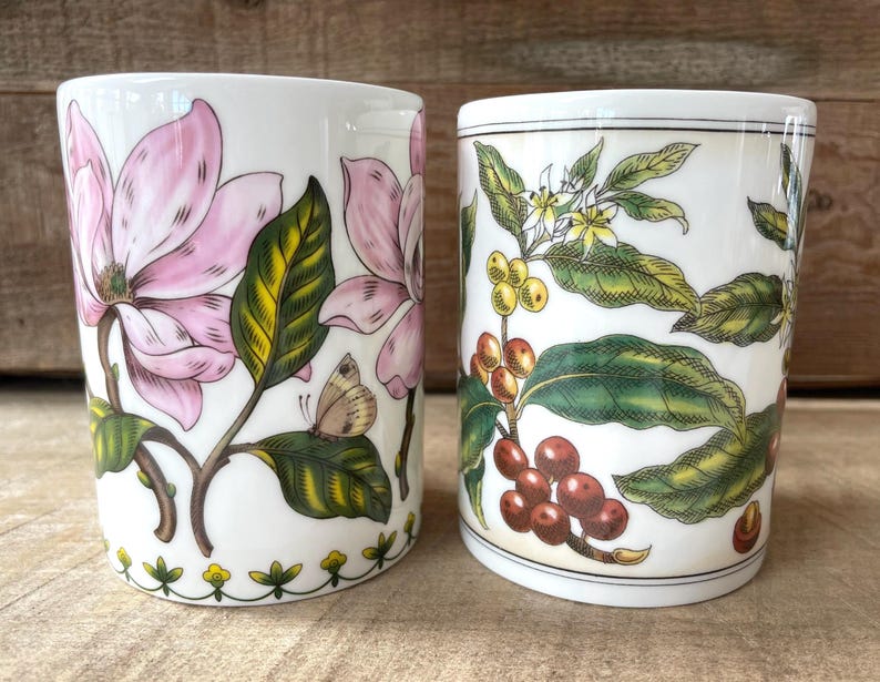 May include: Two white ceramic mugs featuring botanical illustrations. The left mug displays large pink magnolia flowers, green leaves, and a small butterfly. The right mug features coffee plant branches with green leaves, white flowers, and red coffee cherries.