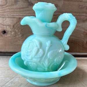 Könnte beinhalten: Ein Set aus einer Kanne und einer Schale aus helltürkisfarbenem Milchglas mit einem erhabenen Rosenmuster. Die Kanne hat einen rosenförmigen Verschluss und einen geschwungenen Griff, während die Schale einen passenden gewellten Rand aufweist.