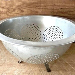 May include: A round, silver-colored metal colander featuring a wide rim and multiple circular sections of small, evenly spaced drainage holes. The bowl-shaped kitchen tool is supported by three short, sturdy metal legs attached to the base.