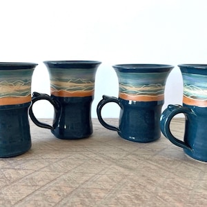 Peut inclure: Quatre tasses en céramique avec une base bleu foncé et un bord évasé. La partie supérieure présente un motif de paysage peint à la main avec des bandes orange, bleu clair et blanc, représentant des collines ou des vagues sur fond bleu.