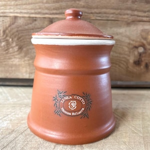May include: A terracotta canister with a matching lid, featuring a white rim and a black printed logo that reads Linea Cotto Mamma Ro Lucca surrounded by leaf illustrations. The cylindrical container has a smooth finish and a flared base.