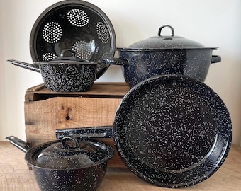 May include: A collection of dark blue enamel cookware with white speckles, including a large colander, a lidded stockpot, a lidded saucepan, and a flat frying pan. Each piece features a smooth, glossy finish and matching dark blue handles.