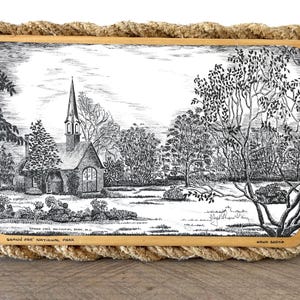 Könnte beinhalten: Eine rechteckige Holztafel mit einer schwarz-weißen Tuschezeichnung einer Kirche im Grand Pre National Park, Nova Scotia. Die Tafel ist von einem dicken, naturfarbenen gedrehten Seil umrandet und enthält den Text Grand Pre National Park, Nova Scotia.