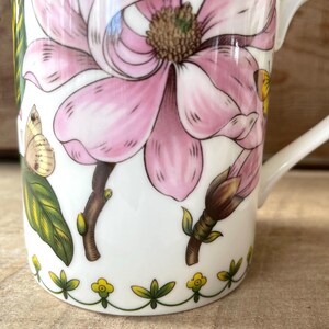May include: A white ceramic mug featuring a large pink magnolia flower, green leaves, and small butterflies. The base of the mug is decorated with a repeating pattern of small yellow flowers and green stems. A white handle is attached to the side.