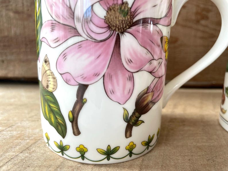 May include: A white ceramic mug featuring a large pink magnolia flower, green leaves, and small butterflies. The base of the mug is decorated with a repeating pattern of small yellow flowers and green stems. A white handle is attached to the side.