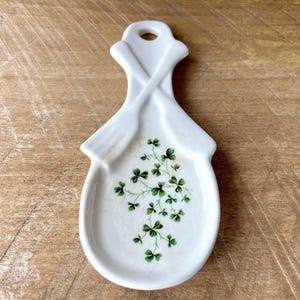 Op de afbeelding: Witte keramische lepelhouder met een klaverontwerp. De lepelhouder heeft een handvat met een gat om op te hangen en een gebogen basis. Het klaverontwerp is groen en is in een cluster op de basis geplaatst.