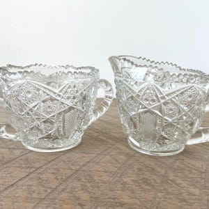 Könnte beinhalten: Ein Set aus Milchkännchen und Zuckerdose aus klarem Pressglas mit einem detaillierten geometrischen Stern- und Rautenmuster. Beide Teile haben gewellte Ränder und robuste, texturierte Griffe mit einer glänzenden Oberfläche.