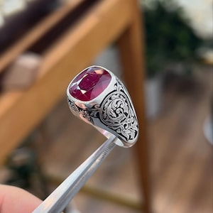 May include: A silver ring with an oval, deep red gemstone. The ring features intricate black floral and scrollwork designs on the band. The gemstone is held in place by silver prongs.