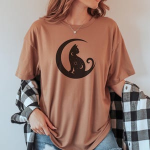 Puede incluir: Camiseta color canela con una silueta de gato negro sobre una luna creciente. El diseño del gato y la luna está centrado en la camiseta. La persona lleva una camisa a cuadros en blanco y negro sobre la camiseta y unos shorts vaqueros azules.