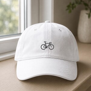 May include: A white baseball cap featuring a small, centred embroidery of a dark grey bicycle. The cap has a structured front panel, a curved brim, and a button on the top. The design is simple, showcasing a minimalist outline of a road bike.