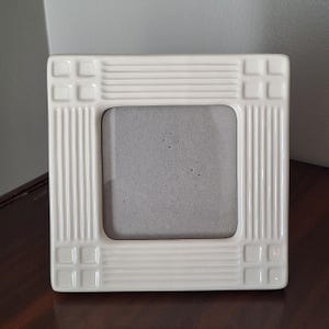 May include: A square, cream-coloured ceramic picture frame. The frame has a rectangular opening for a photo and is decorated with vertical lines and square accents. The frame is on a dark brown surface.