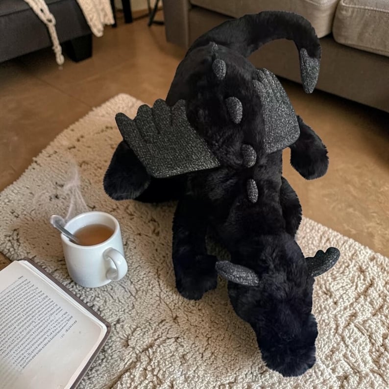 May include: A plush black dragon toy with gray accents on its wings, horns, and back. The dragon is lying on a cream-colored rug. A white mug of tea and an open book are nearby.