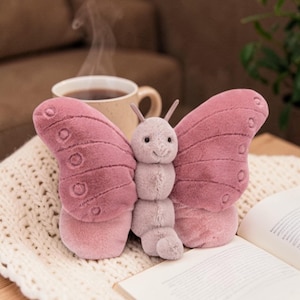 May include: A plush butterfly toy with pink wings and a light purple body. The wings have a textured pattern with circular details. The toy is sitting on a white knitted blanket next to an open book and a cup of coffee.