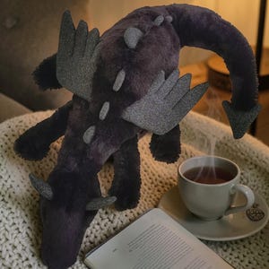 May include: A plush dragon toy in shades of purple and gray, with wings and horns. The dragon is lying on a cream-colored knitted blanket, next to a cup of tea and an open book. The dragon's tail curves downward.
