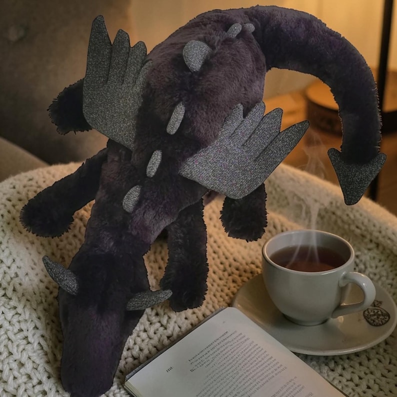 May include: A plush dragon toy in shades of purple and gray, with wings and horns. The dragon is lying on a cream-colored knitted blanket, next to a cup of tea and an open book. The dragon's tail curves downward.