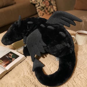 May include: A plush black dragon toy with detailed wings, horns, and a curved tail. The dragon is lying on a soft, textured surface, with a book and a mug of tea in the background.