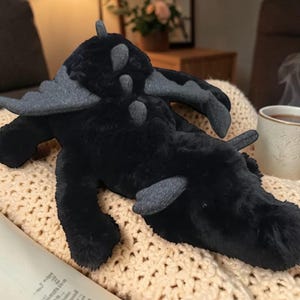 May include: A plush black dragon toy with gray accents on its wings and back. The dragon is lying on a cream-colored knitted blanket. The toy has a soft, furry texture and a whimsical design.