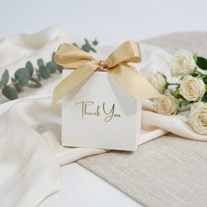 Könnte beinhalten: Eine kleine, weiße Geschenkbox, gebunden mit einer goldenen Satin-Schleife. Die Worte "Thank You" sind in goldener Schrift auf die Vorderseite der Box gedruckt. Die Box befindet sich auf einem cremefarbenen Stoff, mit weißen Rosen und Grünzeug im Hintergrund.