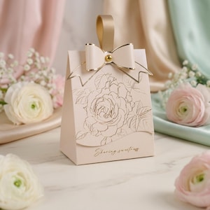 Op de afbeelding: Een lichtroze cadeauzakje met een gouden strik en handvat. De tas heeft een gouden rozenillustratie en de tekst "Sharing sweetness". De tas is omringd door bloemen.
