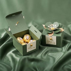 Puede incluir: Dos cajas de regalo verdes sobre una superficie de satén verde. Una caja está abierta, revelando bombones dorados. La otra caja está cerrada y decorada con una flor de papel. Ambas cajas tienen la etiqueta "Joyful Candy".