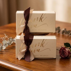 Pode incluir: Duas caixas de presente creme empilhadas, cada uma com a inscrição dourada "Thank You" e "Best Wishes". Elas são amarradas com uma fita marrom escura e creme. Uma rosa e vegetação estão no fundo, em uma superfície de madeira.