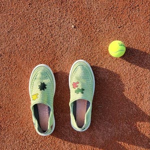 May include: A pair of light green slip-on shoes with floral and geometric designs, next to a yellow tennis ball, all on a reddish-brown surface. The shoes have white soles and are decorated with colorful patches.