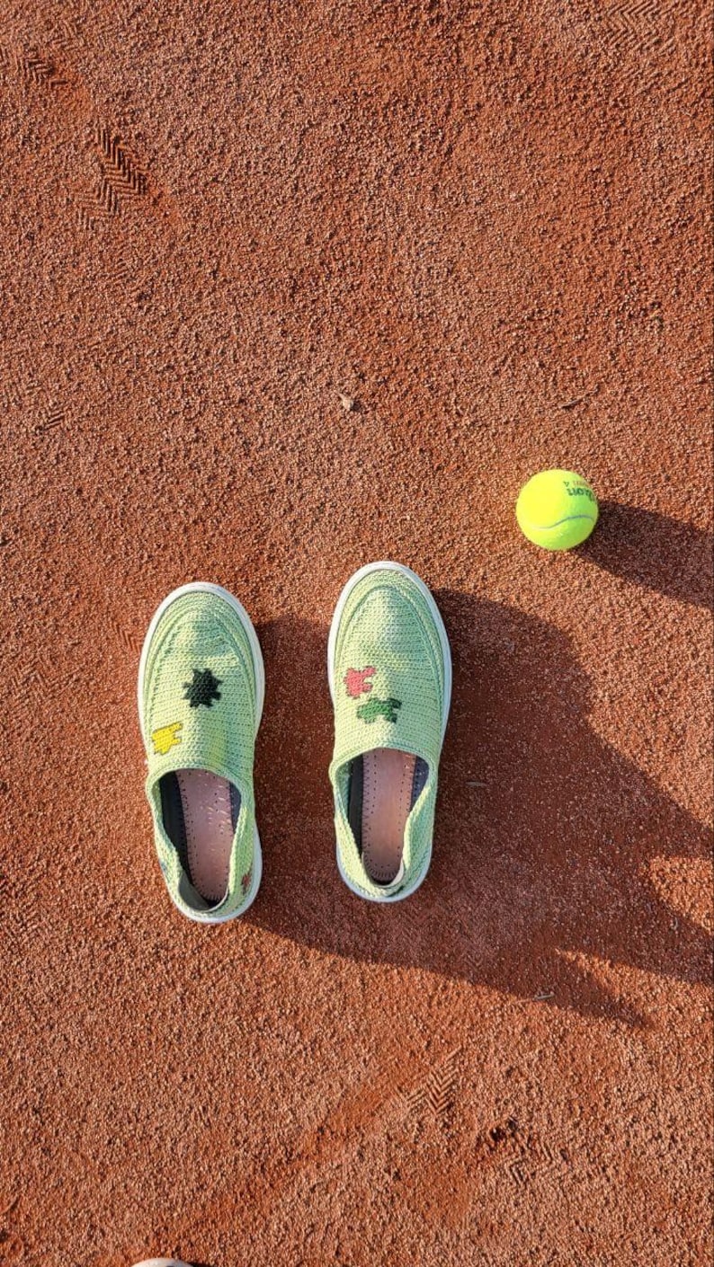 May include: A pair of light green slip-on shoes with floral and geometric designs, next to a yellow tennis ball, all on a reddish-brown surface. The shoes have white soles and are decorated with colorful patches.