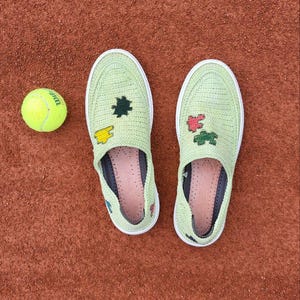 May include: A pair of light green slip-on shoes with white soles and decorative patches, next to a yellow tennis ball. The shoes are on a reddish-brown surface, possibly a tennis court.