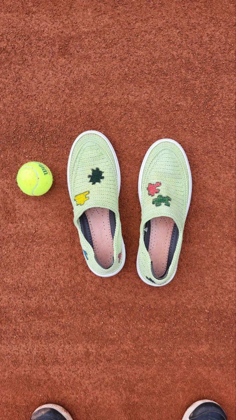 May include: A pair of light green slip-on shoes with white soles and decorative patches, next to a yellow tennis ball. The shoes are on a reddish-brown surface, possibly a tennis court.