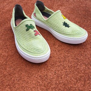 May include: Light green slip-on shoes with white soles. The shoes have a knitted texture and are decorated with embroidered floral designs in various colors. A tennis ball rests nearby on a red surface.
