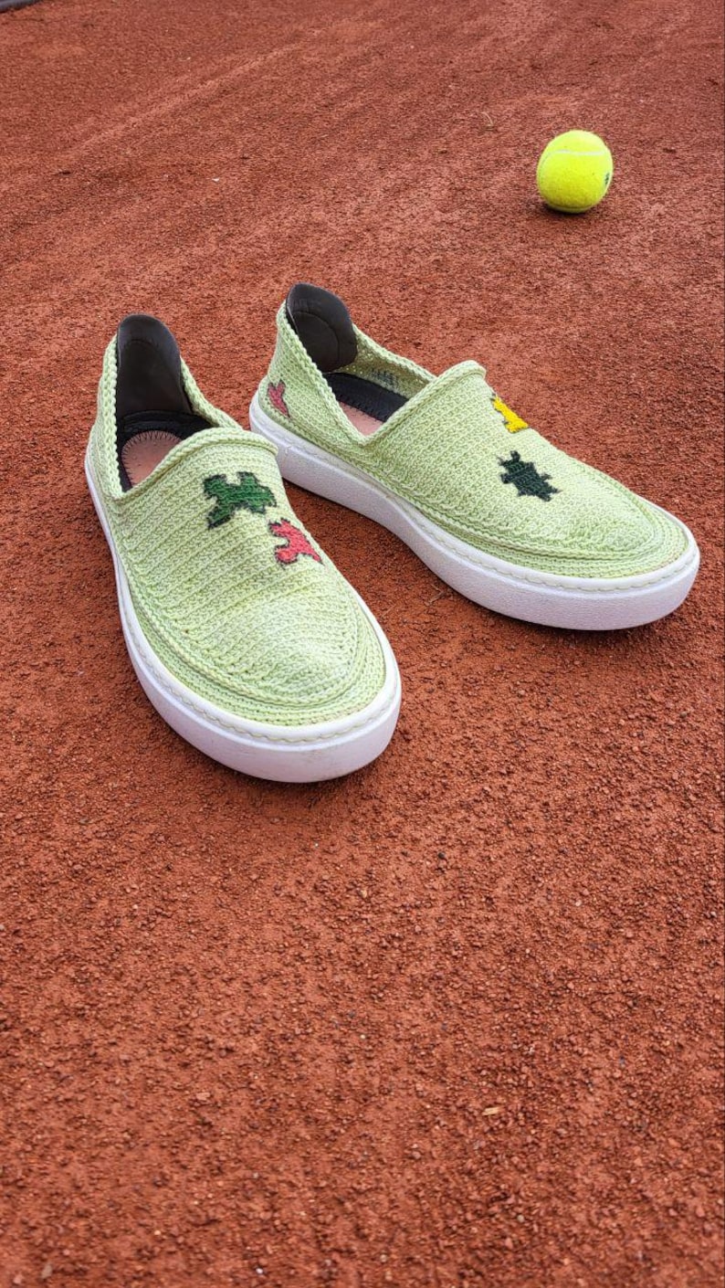 May include: Light green slip-on shoes with white soles. The shoes have a knitted texture and are decorated with embroidered floral designs in various colors. A tennis ball rests nearby on a red surface.