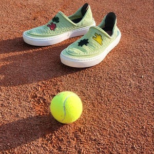 May include: A pair of light green crocheted slip-on shoes with white soles and decorative floral accents, next to a yellow tennis ball. The shoes are on a reddish-brown surface, likely a tennis court.