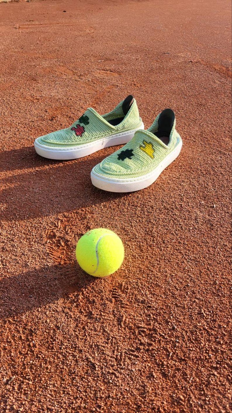 May include: A pair of light green crocheted slip-on shoes with white soles and decorative floral accents, next to a yellow tennis ball. The shoes are on a reddish-brown surface, likely a tennis court.