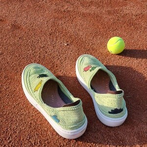 May include: A pair of light green crochet slip-on shoes with white soles, decorated with small embroidered details. A yellow tennis ball rests nearby on a reddish-brown surface. The shoes have a casual, handmade aesthetic.