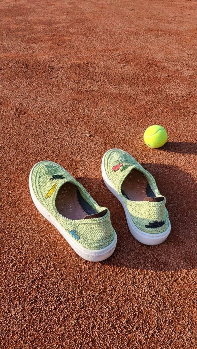 May include: A pair of light green crochet slip-on shoes with white soles, decorated with small embroidered details. A yellow tennis ball rests nearby on a reddish-brown surface. The shoes have a casual, handmade aesthetic.