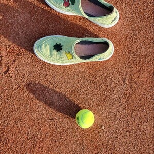 May include: Two light green crochet slip-on shoes with white soles and embroidered details, next to a yellow tennis ball, all on a reddish-brown surface. The shoes have black and pink accents.