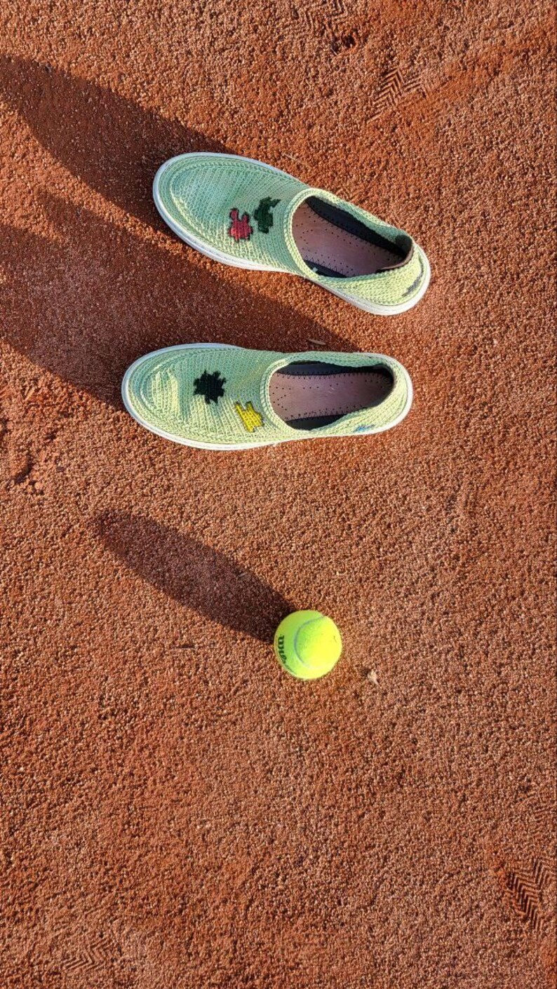 May include: Two light green crochet slip-on shoes with white soles and embroidered details, next to a yellow tennis ball, all on a reddish-brown surface. The shoes have black and pink accents.
