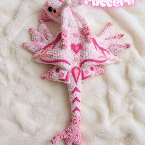 May include: A handmade pink and white crochet creature with a heart design on its body. The creature has multiple wing-like appendages and the text "Crochet Pattern" in a pink, glowing font.