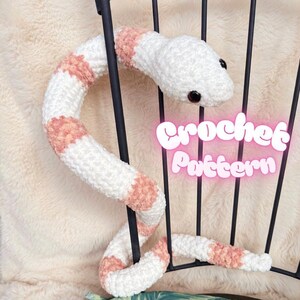 May include: A white and peach crochet snake with black eyes is draped over a black metal frame. The snake has peach-colored bands along its body. The words "Crochet Pattern" are in pink neon text.