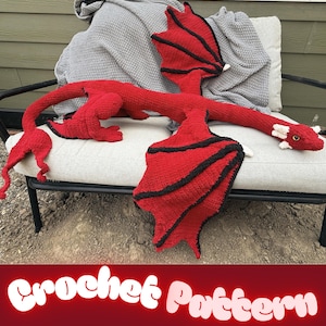May include: A large, handmade crochet dragon toy featuring a vibrant red body with black-trimmed wings and white accents on the head. The dragon has a long, slender neck, a tail with a pointed tip, and textured yarn construction throughout.
