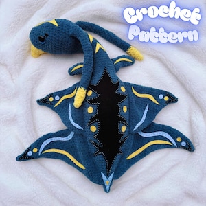 Puede incluir: Una criatura marina de ganchillo hecha a mano en tonos azules, amarillos y negros. El peluche presenta un cuello largo y un cuerpo en forma de estrella, con el texto "Crochet Pattern". Está sobre una superficie blanca y esponjosa.