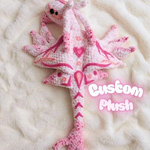 May include: A handmade pink and white crochet plush toy with a heart design. The plush has a long tail and wings, with the text "Custom Plush" in a glowing pink font.