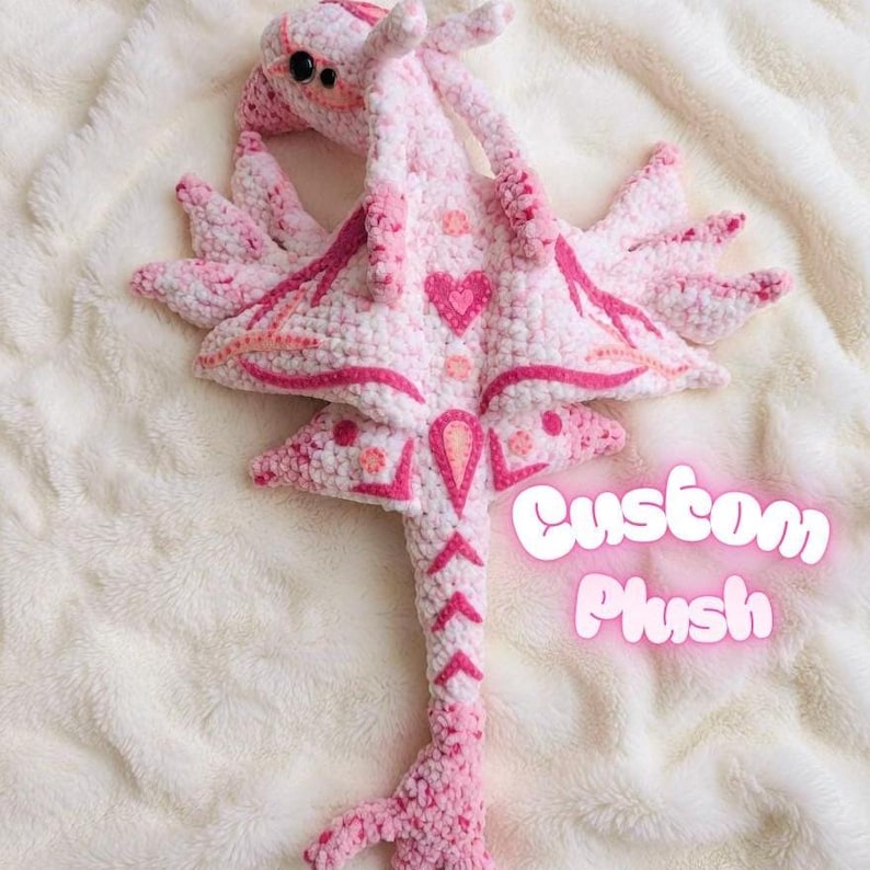 May include: A handmade pink and white crochet plush toy with a heart design. The plush has a long tail and wings, with the text "Custom Plush" in a glowing pink font.