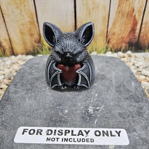 May include: A small, dark gray bat figurine with textured fur and large ears, holding a smooth, deep red heart against its chest. The bat has closed eyes and is positioned on a gray stone base with a white label reading FOR DISPLAY ONLY NOT INCLUDED.